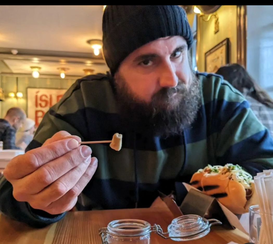 An image of a bearded man in a winter hat, offering a small piece of food to the camera. The food is hakarl, a fermented shark dish he enjoyed in Iceland.